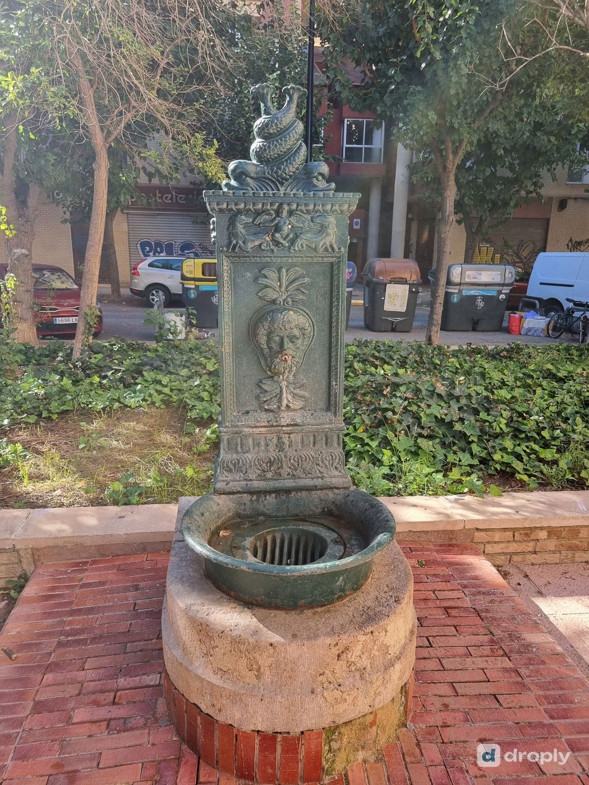 Nahaufnahme eines grünen Trinkbrunnens aus Gusseisen in Benimaclet, Valencia, mit dem bärtigen Gesicht und dem Wasserspeier im Mund, der Palmette darüber und zwei verschlungenen Delfinen obendrauf