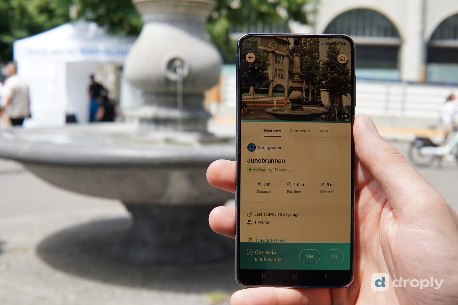 Une main tient un téléphone devant le Junobrunnen à Zurich, l'appli droply affiche la fontaine comme « Coule » avec la question du check-in et les boutons Oui et Non