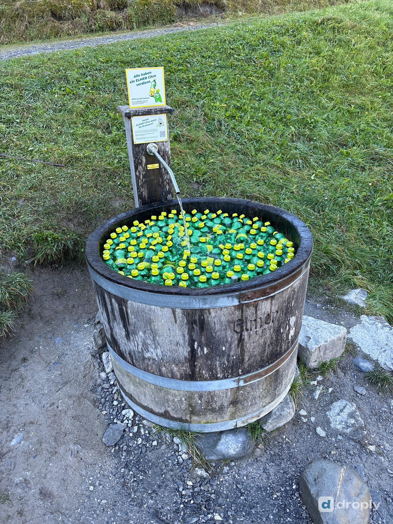 Ein Holzfassbrunnen voller grüner Elmer-Citro-Flaschen, die im laufenden Wasser kühlen, oberhalb von Elm im Glarnerland