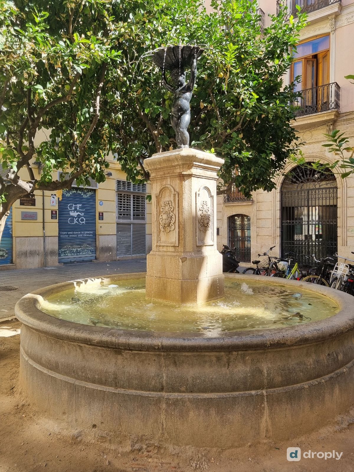 Die Font del Negret in Valencia, eine Kinderfigur, die eine Muschel hochhält, auf einem Steinsockel mit Gesichtern an den Seiten, über einem runden Steinbecken