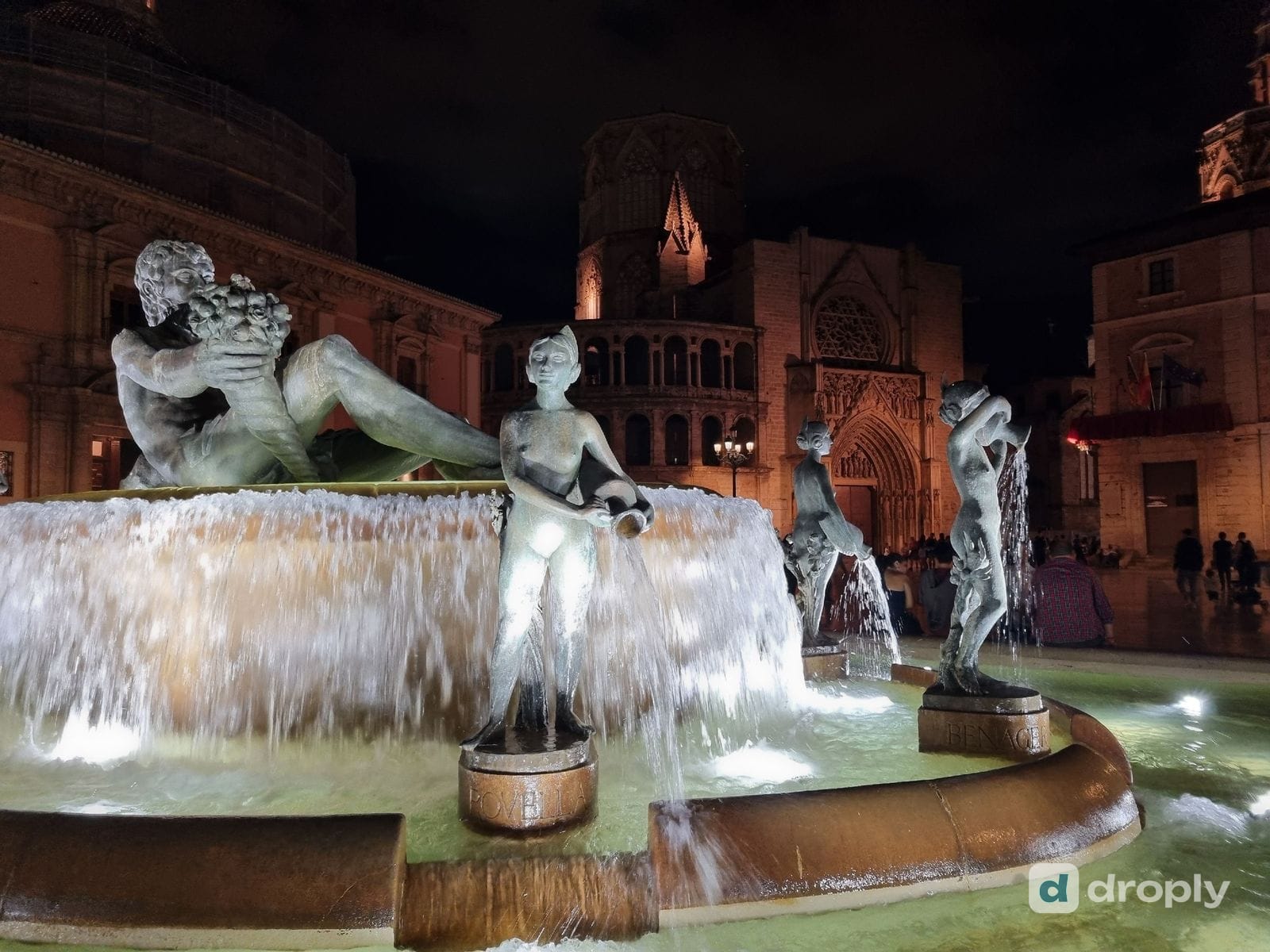 Der Turia-Brunnen auf der Plaça de la Mare de Déu bei Nacht, mit dem bronzenen Flussgott mit Füllhorn und den Figuren Rovella und Benager, die Wasser aus Krügen giessen, dahinter das gotische Apostelportal der Kathedrale von Valencia