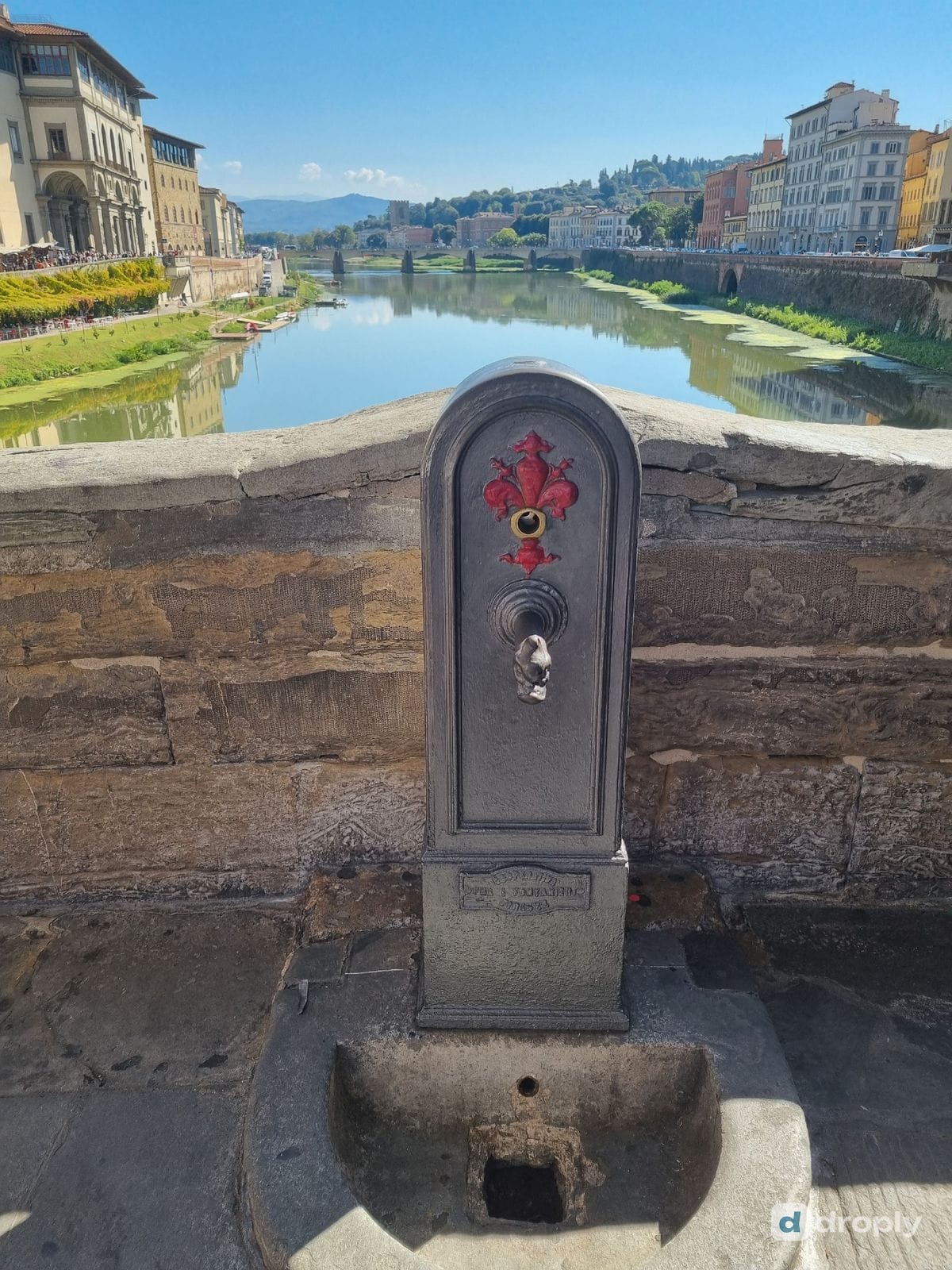 A simple iron drinking tap on a stone embankment along the Arno river in Florence, with the river and historic buildings behind it