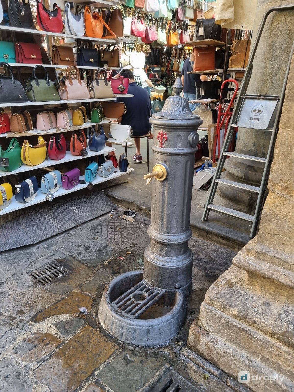 A drinking tap in Florence standing next to leather goods stalls at the San Lorenzo market, with shoppers in the background