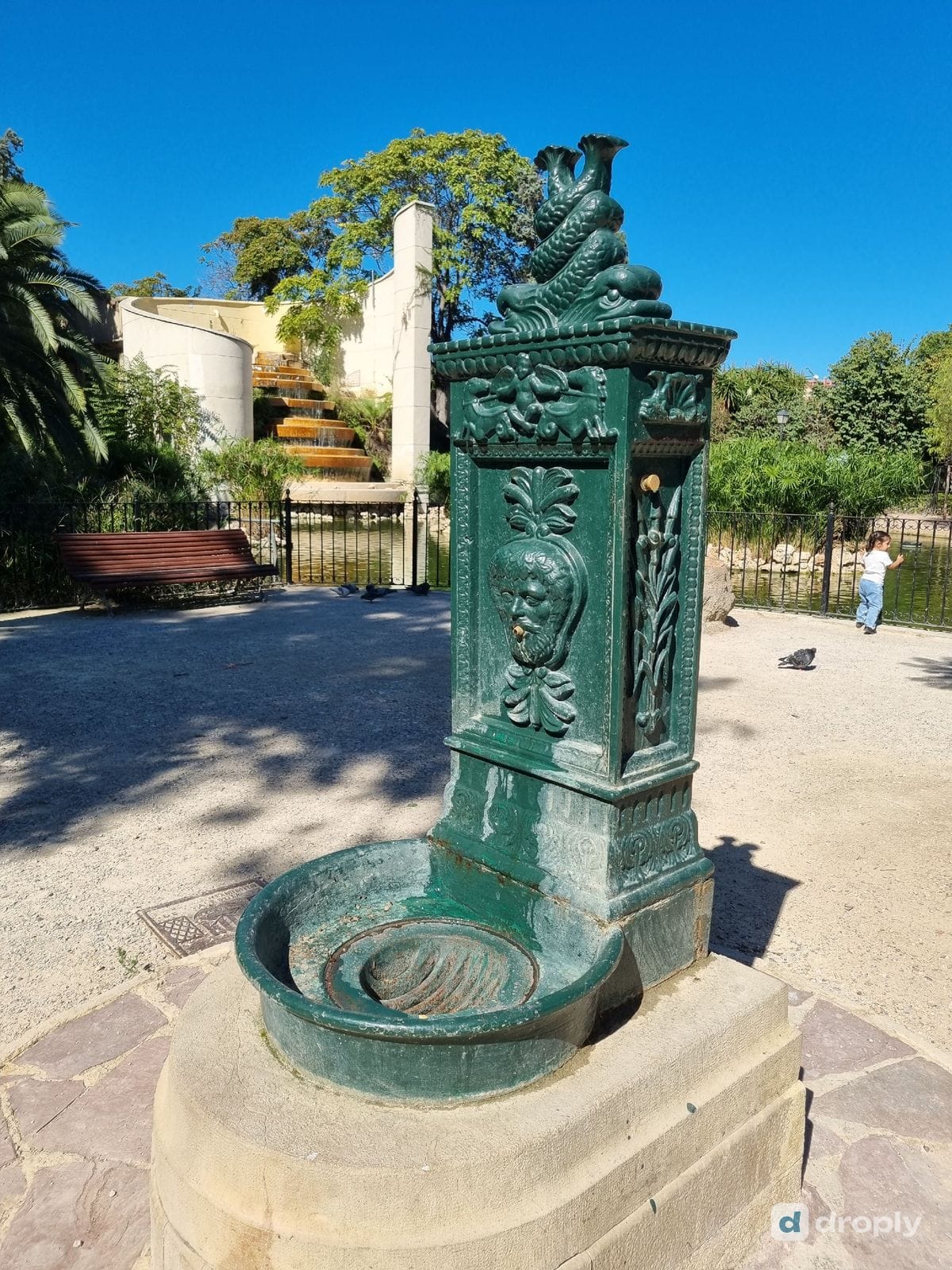 Ein grüner Trinkbrunnen aus Gusseisen mit einem bärtigen Gesicht als Wasserspeier, einem Puttenfries und zwei ineinander verschlungenen Delfinen obendrauf, neben einem Teich im Turia-Park in Valencia