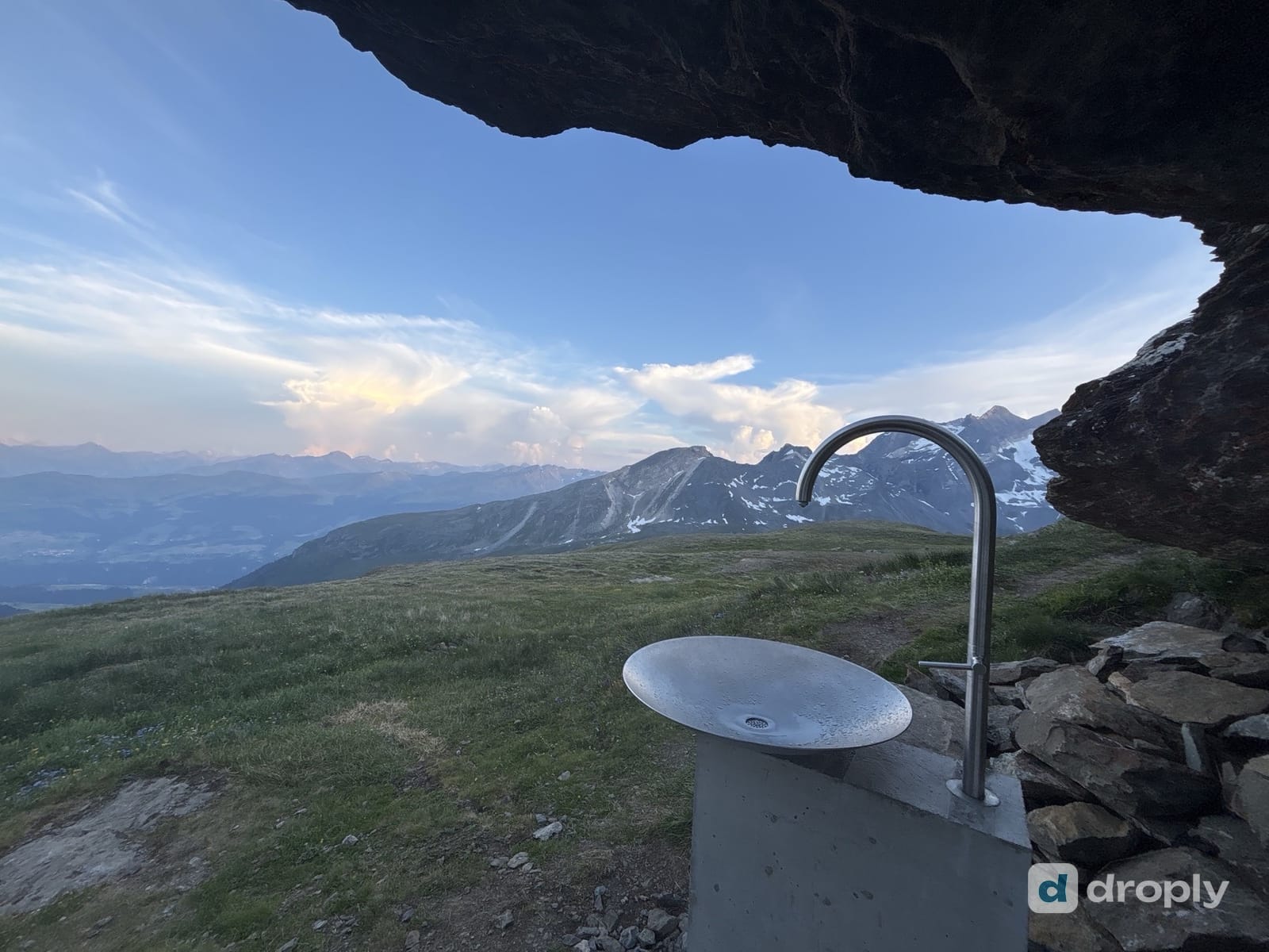 Ein Edelstahlbrunnen unter einem Felsvorsprung oberhalb von Breil/Brigels in Graubünden, im Hintergrund schneegestreifte Gipfel