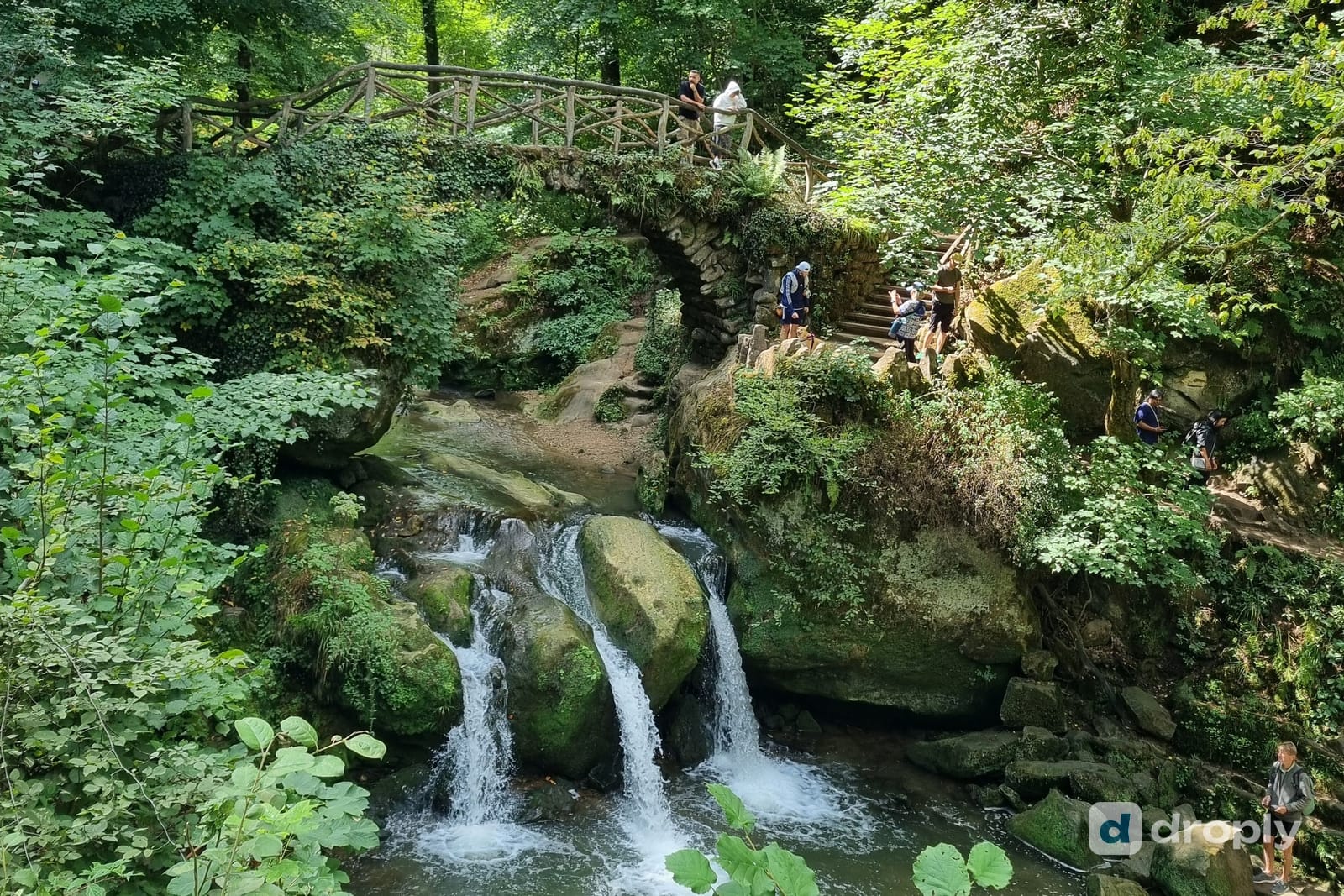 Der Wasserfall Schéissendëmpel mit Steinbrücke und Holztreppe auf dem Mullerthal Trail in Luxemburg