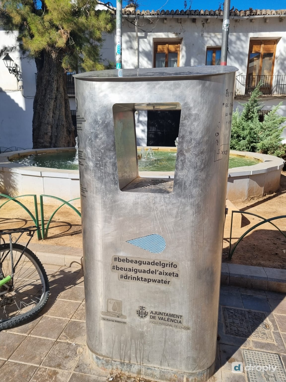 Ein PUSDAR-Nachfüllspender aus Edelstahl in Valencia mit den Hashtags #bebeaguadelgrifo, #beuaiguadelaixeta und #drinktapwater und dem Logo des Ajuntament de València, vor einem kleinen Platz mit einem Brunnen