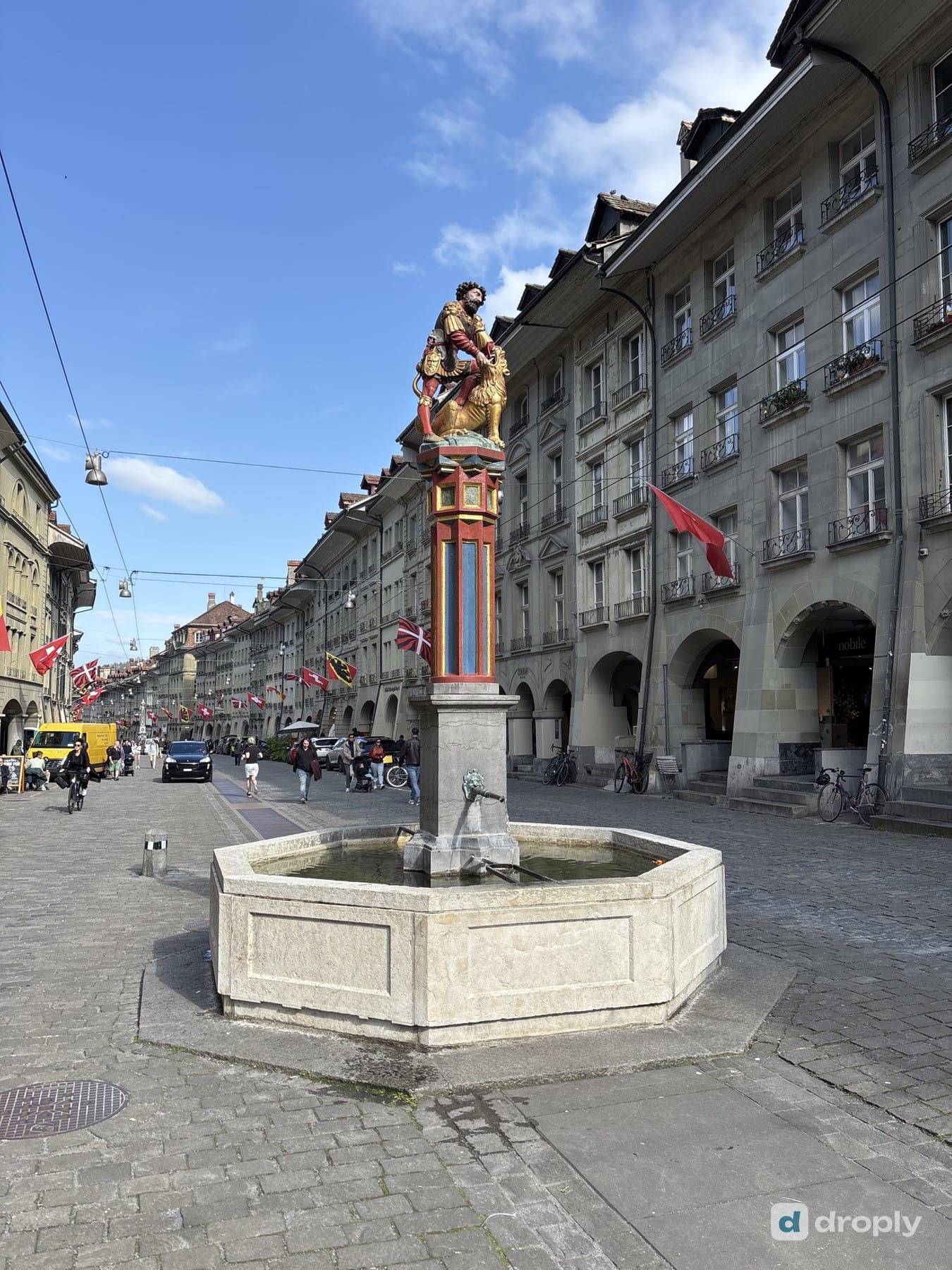 The Simsonbrunnen on Kramgasse in Bern under a blue sky, with Swiss flags on the facades