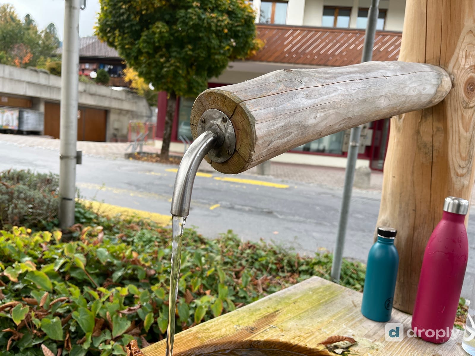 Ein hölzerner Dorfbrunnen läuft in einen Trog, daneben stehen eine blaue und eine pinke Trinkflasche, in Mannried bei Zweisimmen